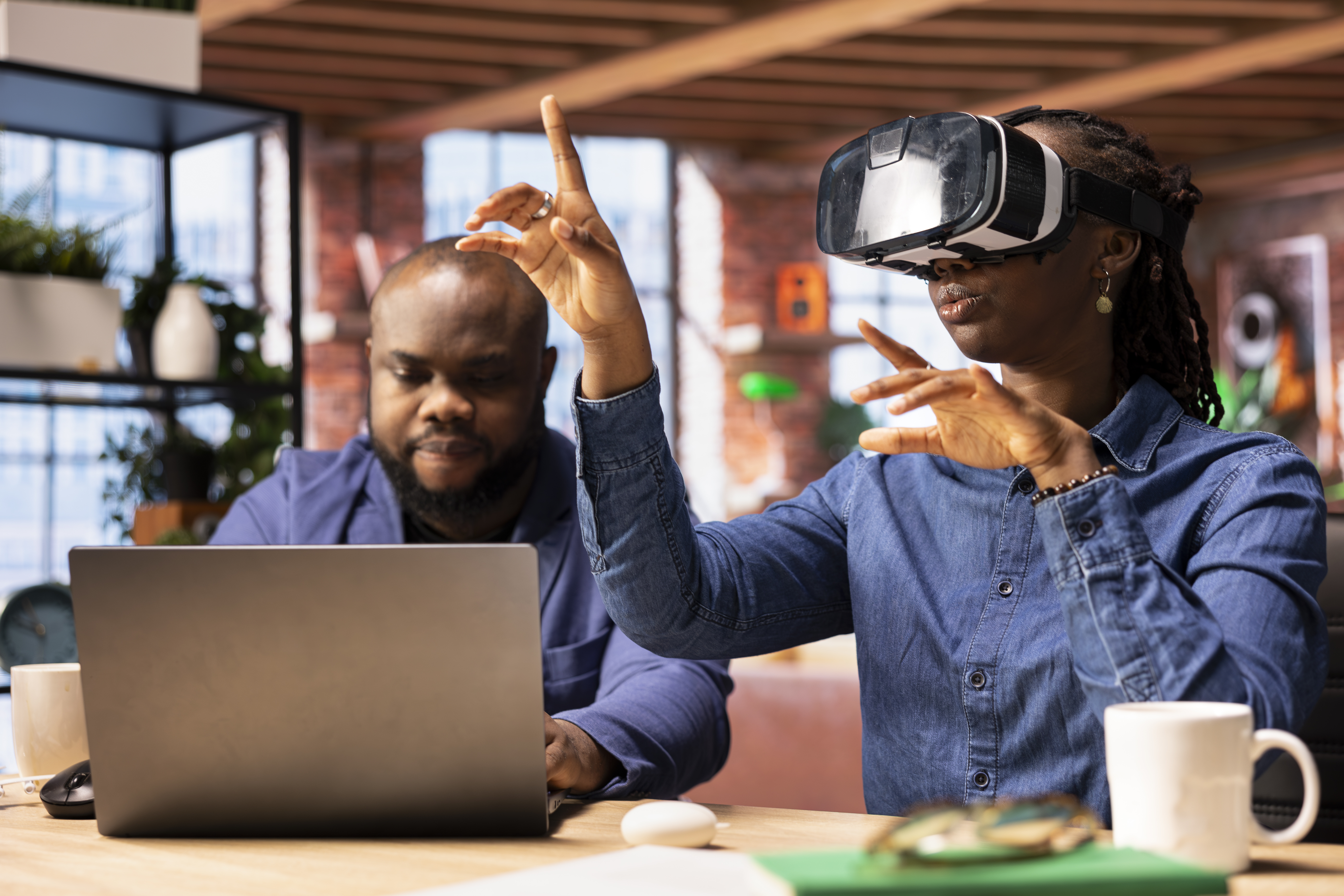 Black girl engages with virtual reality while her partner works on a laptop at the desk, exploring AI and interactive technology in a modern home office setting. 3D simulation with gadgets.